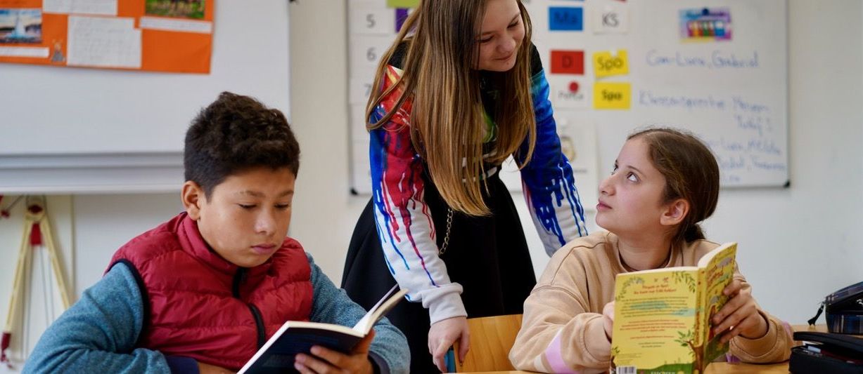 Ein Schüler liest in einem Buch, daneben unterhalten sich zwei Schülerinnen