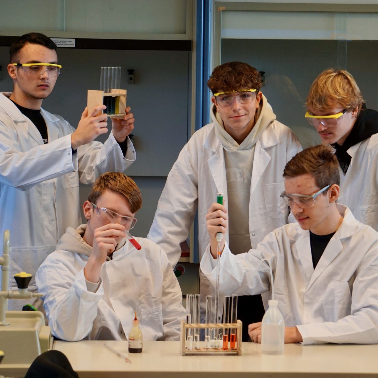 Eine Schülergruppe in weißen Kitteln und mit Schutzbrillen bei einem Experiment im naturwissenschaftlichen Unterricht